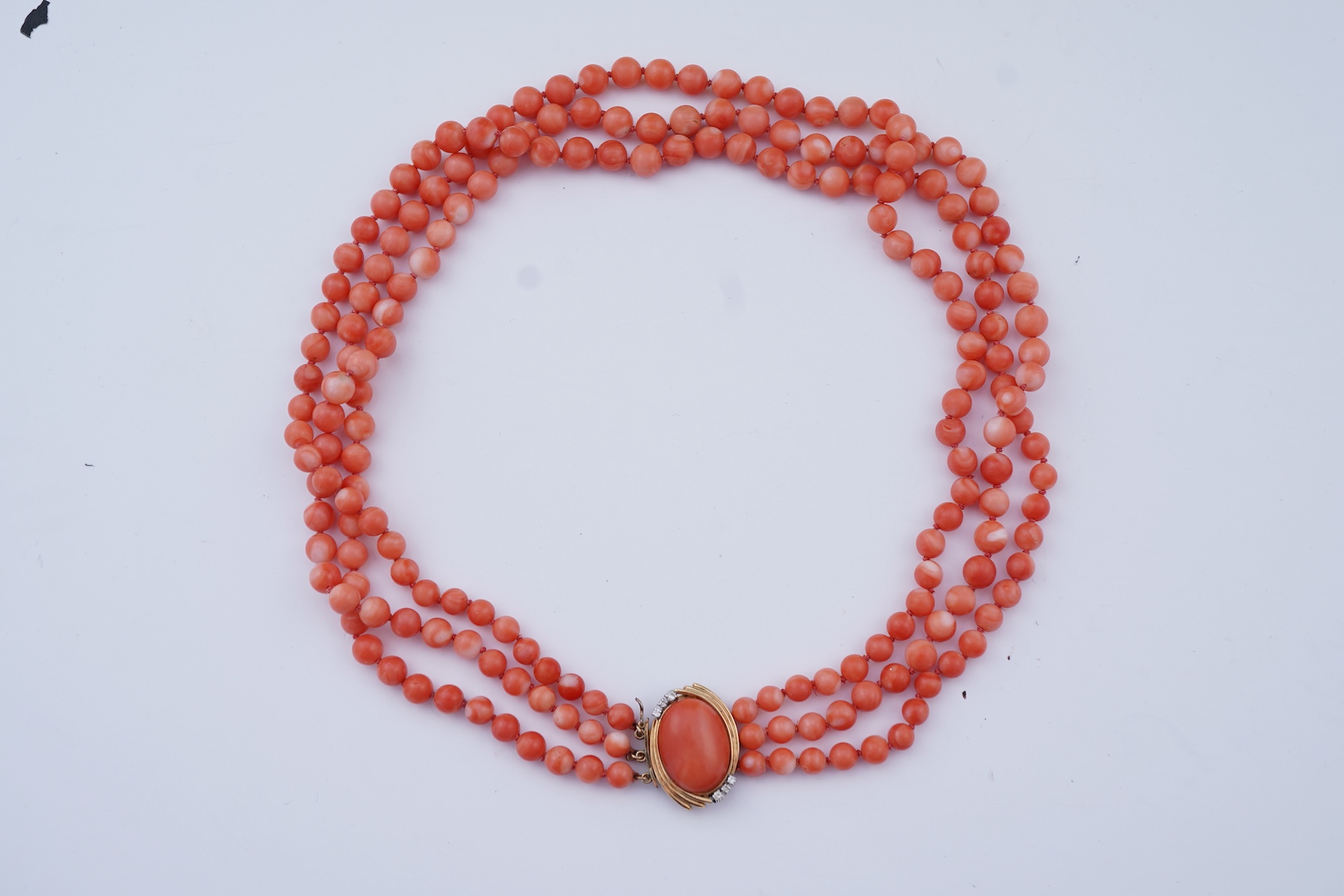A coral necklace, composed of three strands of coral beads measuring approximately 5.1-6.6mm diameter, to a gold clasp set with a coral cabochon and brilliant-cut diamonds, mounted in gold, length 45.5cm, stamped 14K, ca